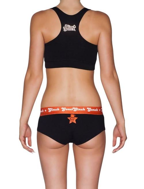 Full back view of a model wearing Ginch Gonch Rock Me Gogo Women's Briefs in black with orange waistband, skull and guitar rockstar print stretch underwear and a black sports bra.
